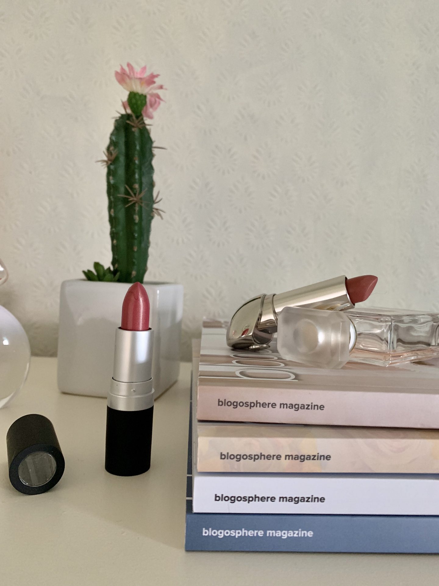 A photo of a dark nude lipstick next to four copies of Blogosphere Magazine which are stacked on top of each other. Another dark nude lipstick in a silver tube is placed on top of the magazines and a fake cactus can be seen in the background
