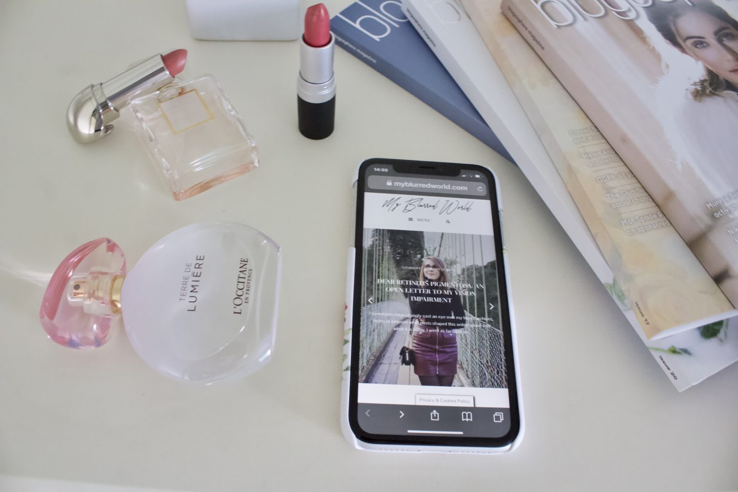 A photo of my blog's homepage displayed on a phone. There are a couple of lipsticks around it, one standing upright whilst the other leans on a small Chanel perfume bottle