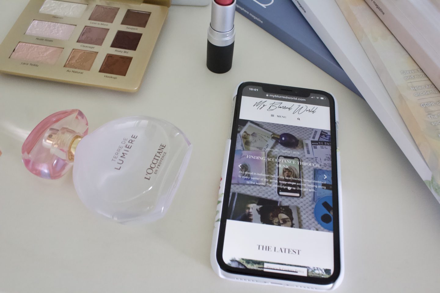 A photo of my blog's homepage displaying on an iPhone, there is a L'occitane perfume beside it, a dark pink lipstick and a small Too Faced eyeshadow palette