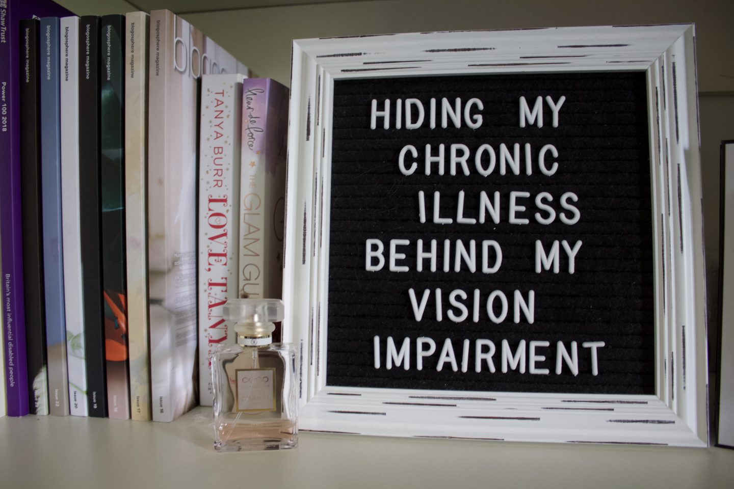 A photo of a vintage white framed black letter board which says hiding my chronic illness behind my vision impairment, a small bottle of Chanel perfume stands upright in the left corner