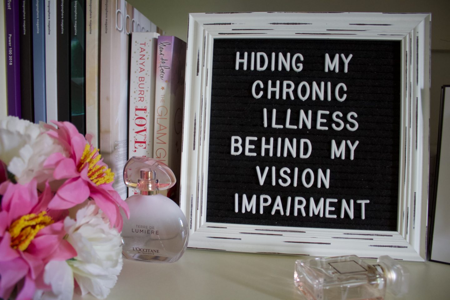 A photo of a white framed black letter board which has the writing Hiding my chronic illness behind my vision impairement in white, it's proppe up beside a few copies of Blogosphere Magazine which are upright along with a couple of other books. There are a couple of perfumes in front of the letter board.