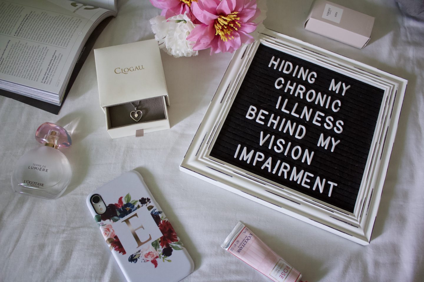 A flatlay photo of a white framed black letter board which says Hiding my chronic illness behind my vision impairment, it's surrounded by a few items such as a white jewellery box, a light pink tube of hand cream and white and pink fake flowers