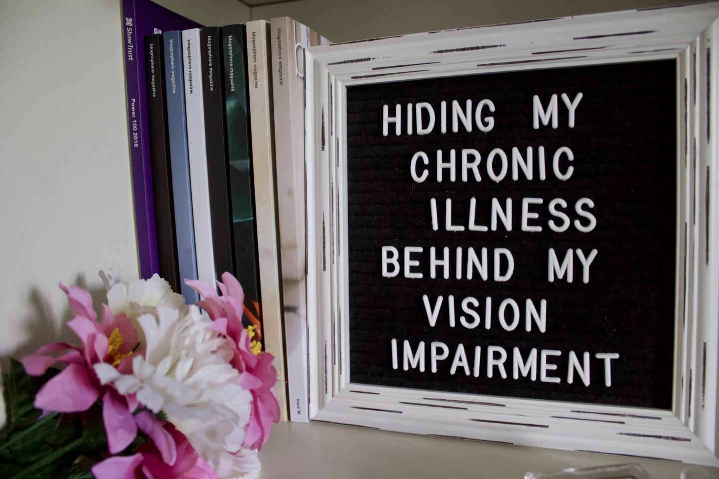 A photo of fake pink and wite flowers next to a white framed black letter board which displays the blog post title. There are a few copies of Blogosphere Magazine standing upright beside the frame on the left