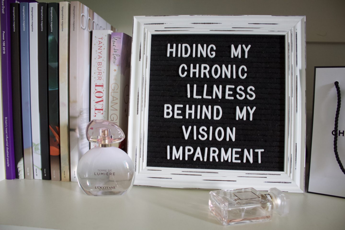 A photo of a white vintage framed black letter board which says Hiding my chronic illness behind my vision impairment. There are a couple of perfumes in front of it, one is upright on the left hand corner whilst the other is horizontal in front of the frame