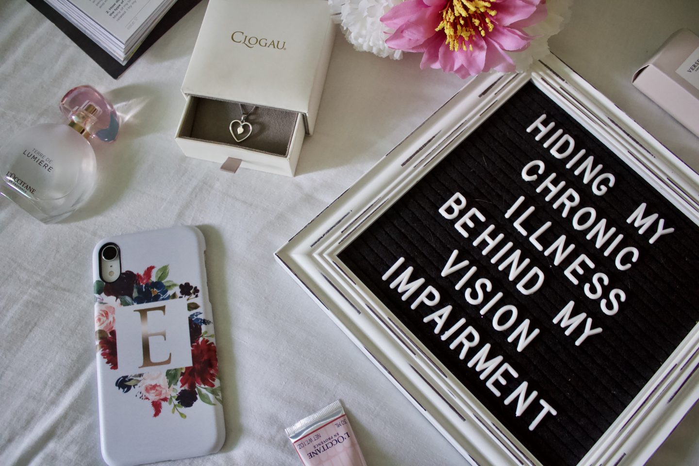 A photo of a L'occitane perfume, a white phone case with the letter E on it in silver and a silver heart shaped necklace surrounding a white framed letter board on a white duvet cover