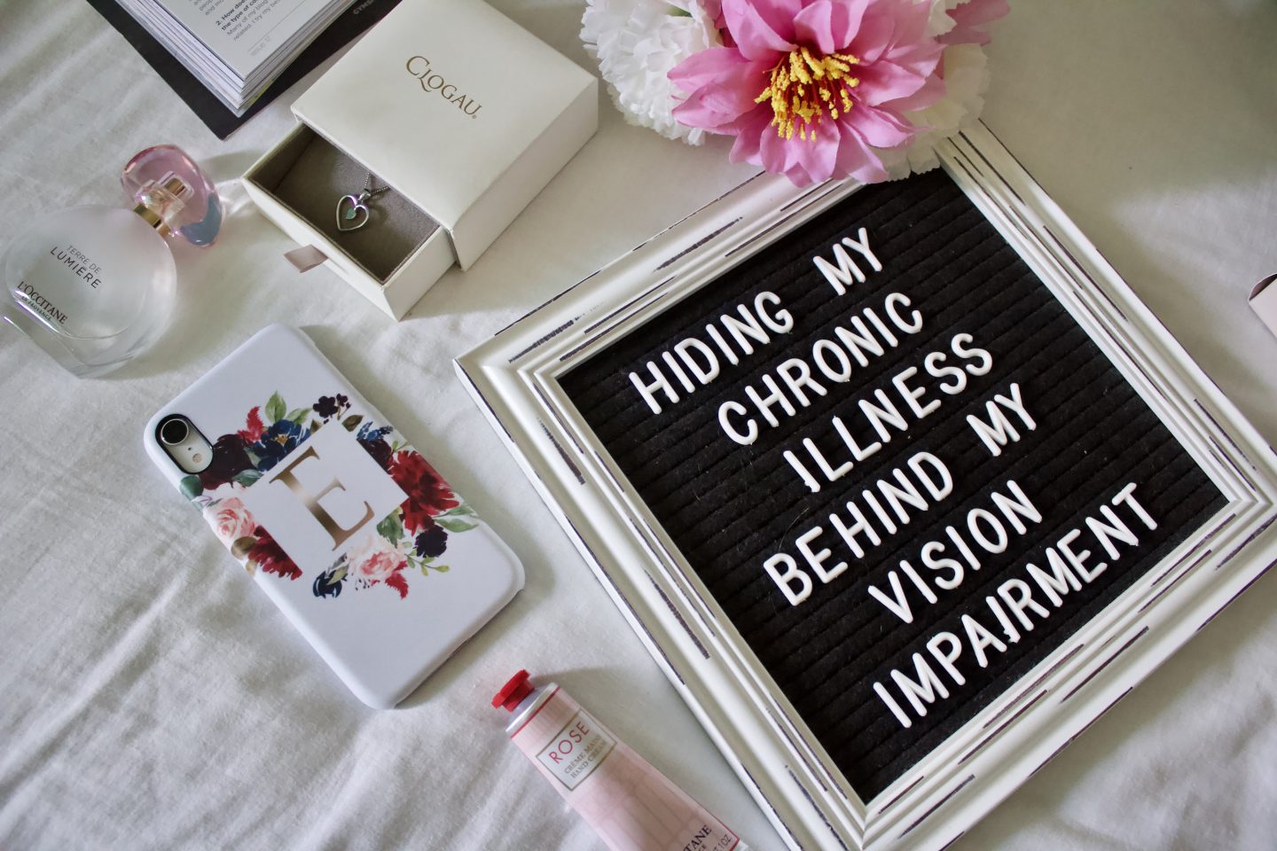 A photo of a white framed felt letter board on a white duvet cover, it's surrounded by various products including some pink and white fake flowers and a circular perfume bottle