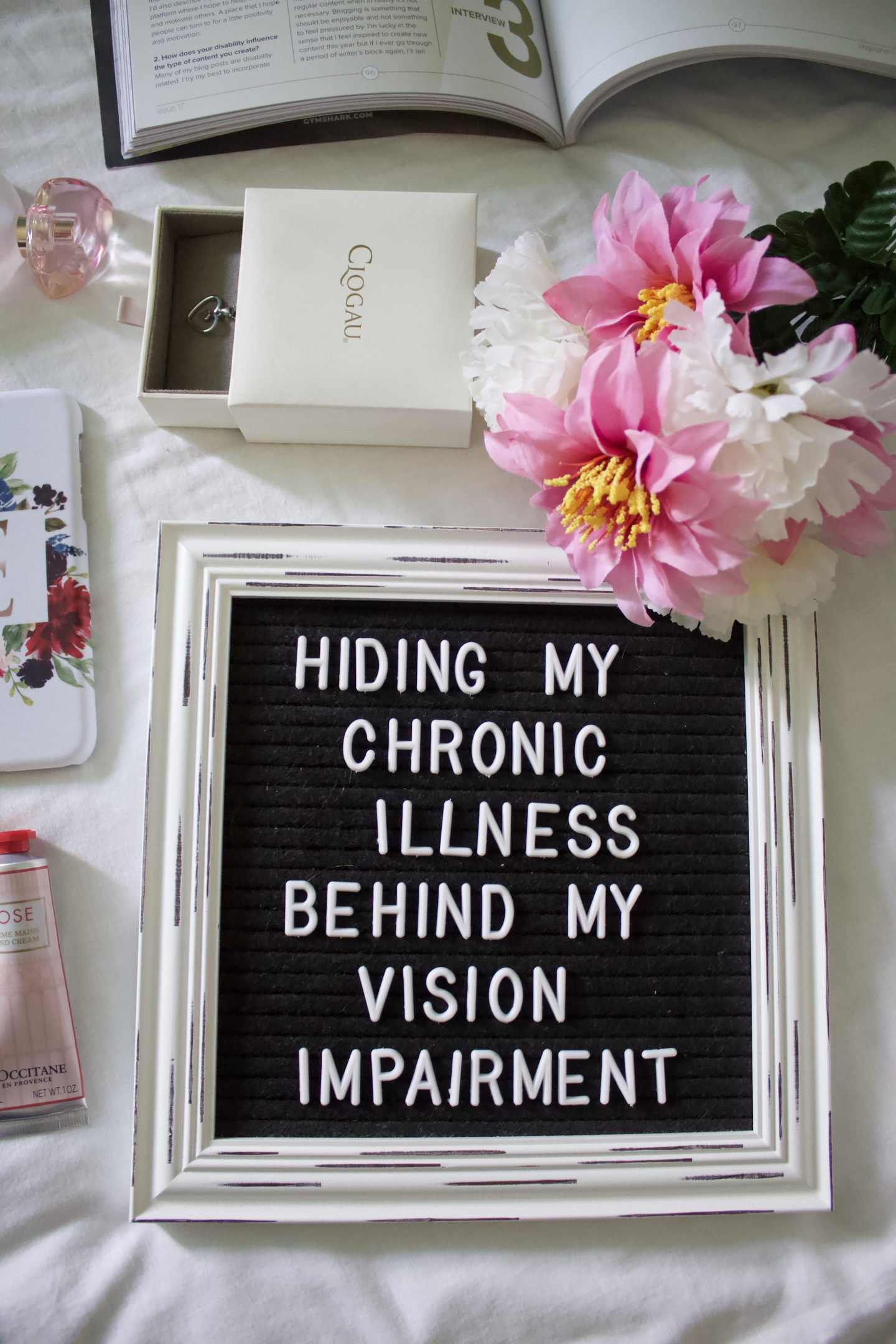 A photo showing the white framed black letter board on a white duvet, it's surrounded by things such as pink and white fake flowers, a white necklace box with a silver heart shaped necklace poking out of it and a light pink coloured tube of hand cream