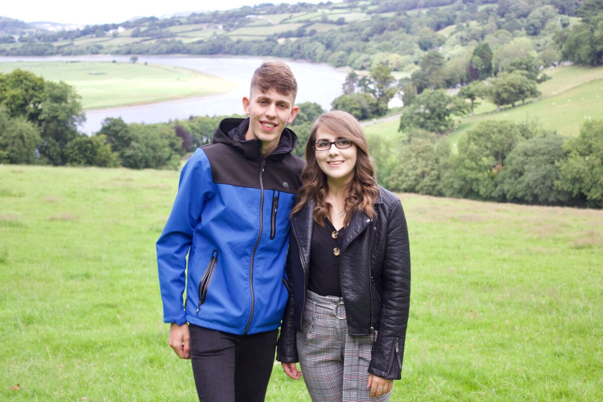 A photo of my brother and I standing in a field which overlooks a view of trees and a river. We're both smiling at the camera