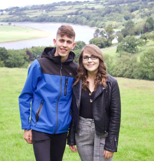 A photo of my brother and I standing in a field which overlooks a view of trees and a river. We're both smiling at the camera