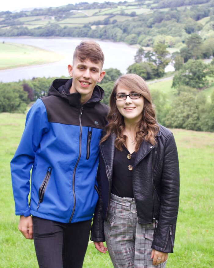 A photo of my brother and I standing in a field which overlooks a view of trees and a river. We're both smiling at the camera