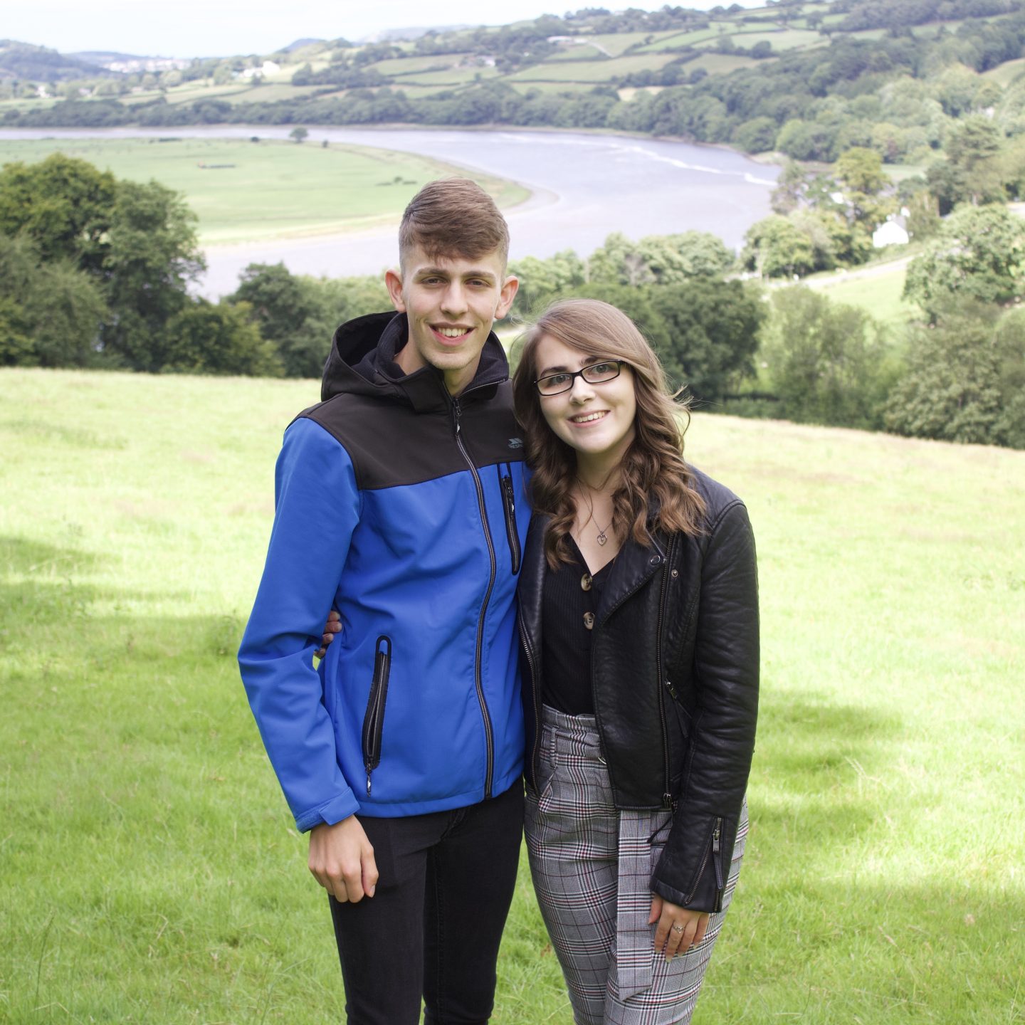 A photo of my brother and I standing in a field which overlooks a view of a river and trees. We've got our arms around each other.