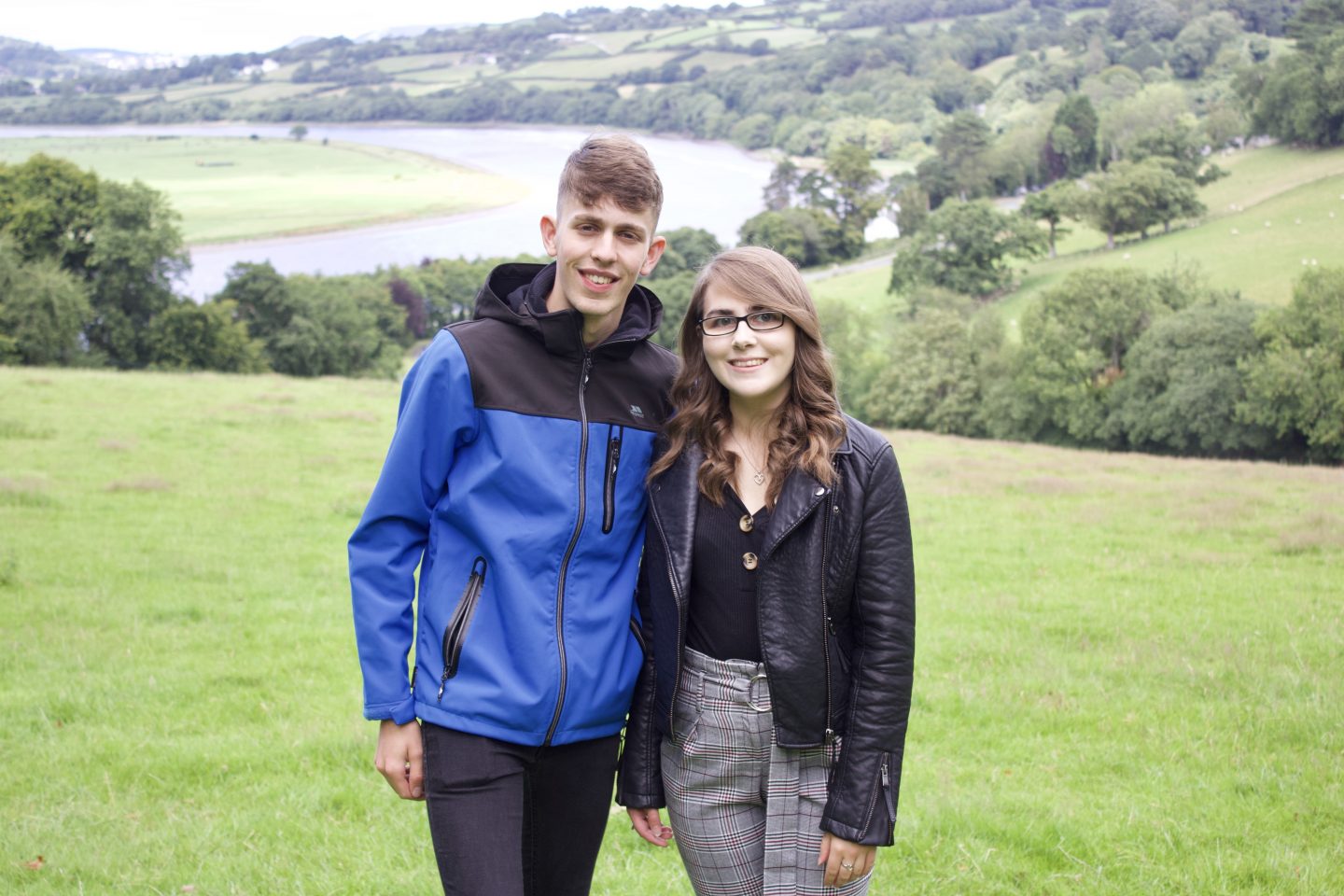 A photo of my brother and I standing in a field which overlooks a view of trees and a river. We're both smiling at the camera
