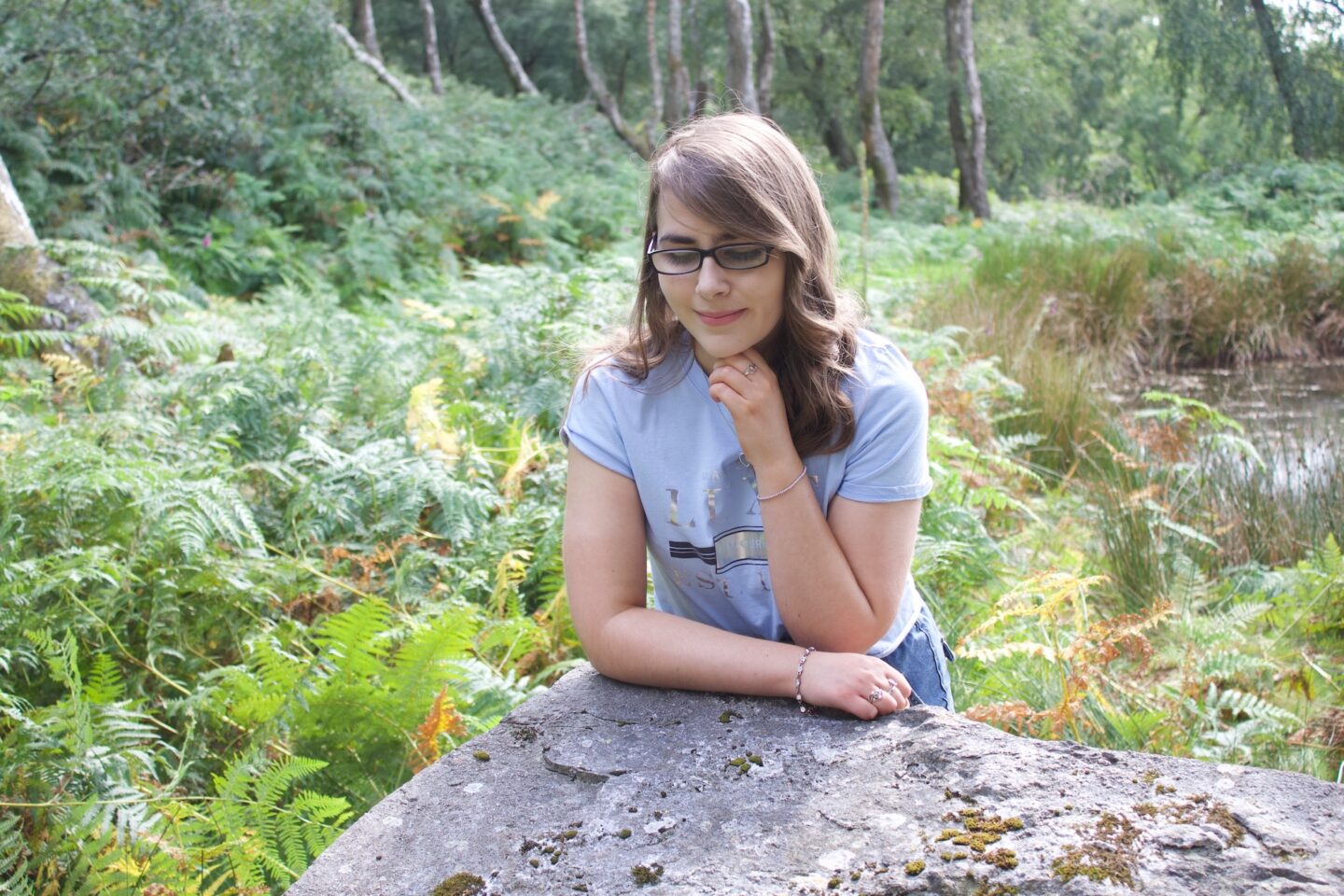 A photo of Elin leaning forward on a big rock, her right arm rests on the rock whilst her left is raise to just underneath her chin. She's looking down and there are trees around her