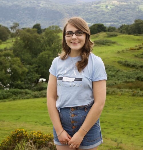 A photo of Elin standing in front of a view of mountains, she wears a light blue cropped t-shirt with high waisted denim shorts