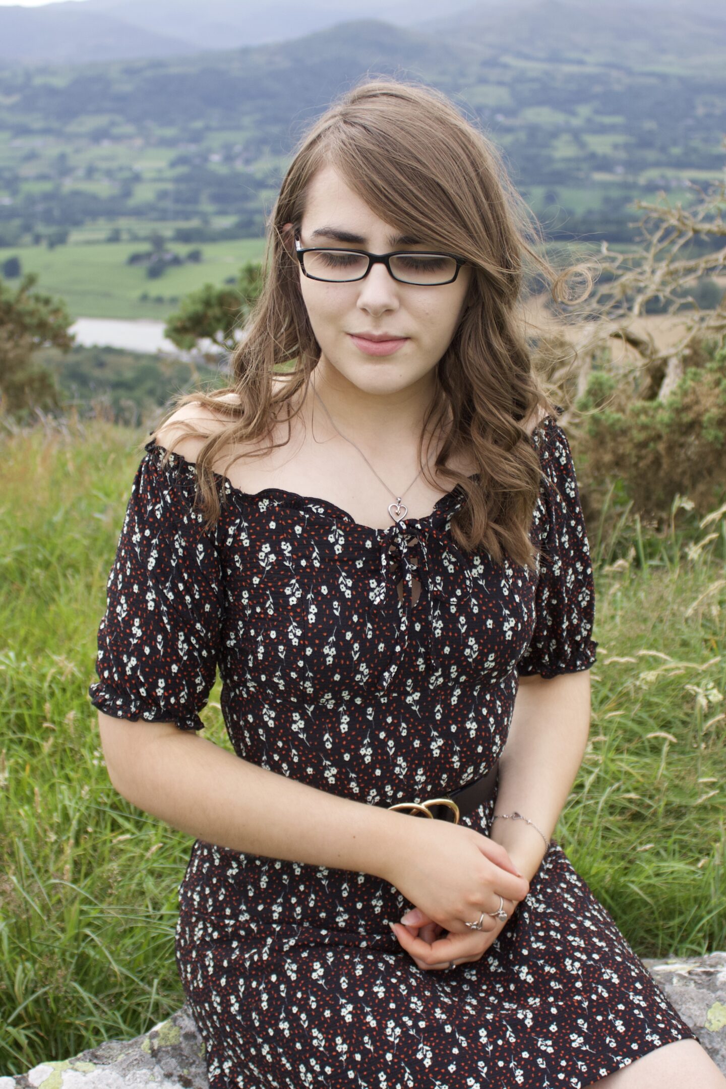 A photo of Elin looking down, she's sitting on a rock with green hills in the background