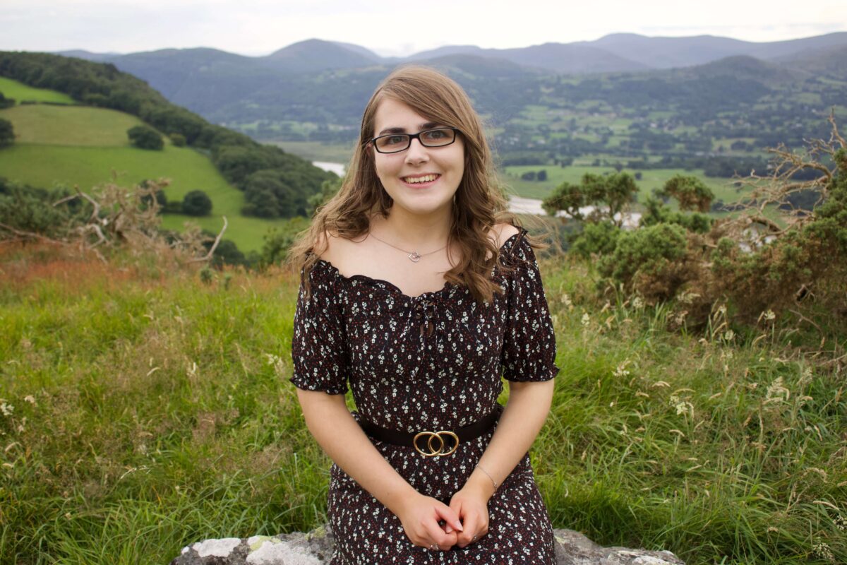 A photo of Elin sitting on a rock with a few of a river and mountains in the background. She's wearing a black bardot floral dress with a black belt which has two circles overlapping in the centre.