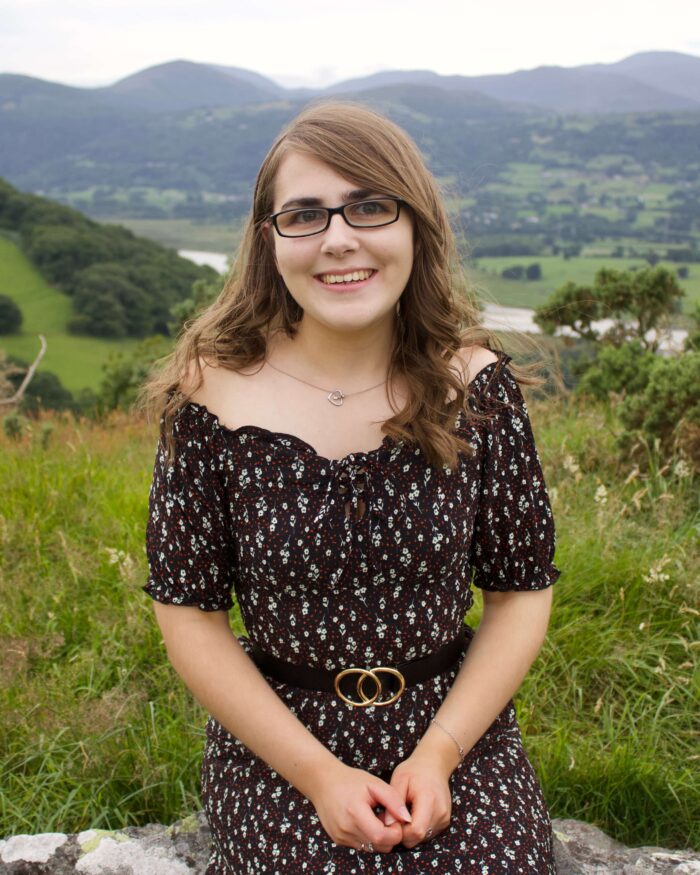 A photo of Elin sitting on a rock with a few of a river and mountains in the background. She's wearing a black bardot floral dress with a black belt which has two circles overlapping in the centre.