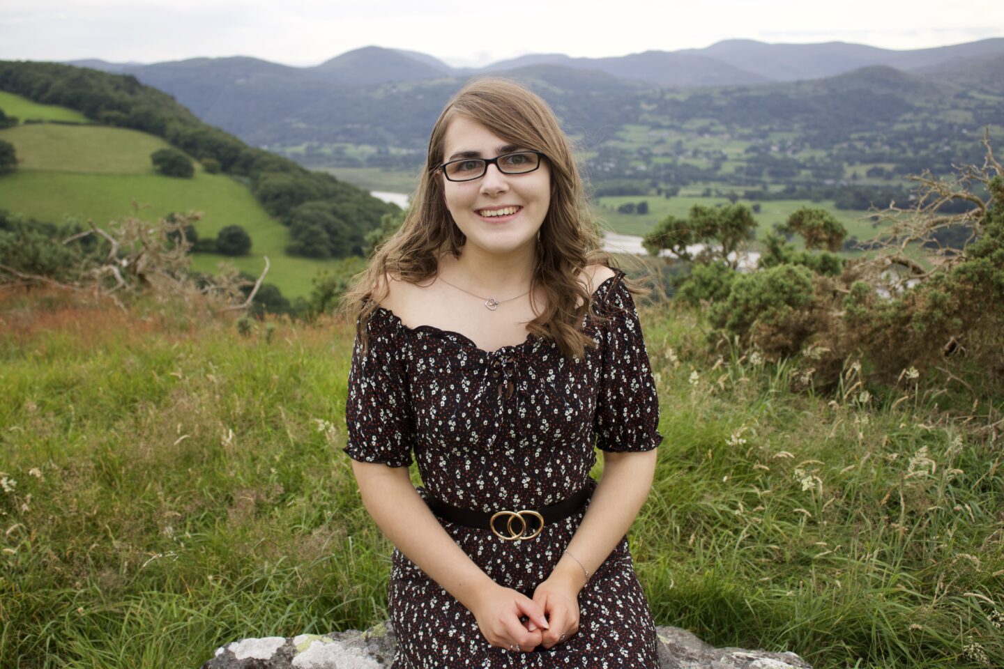 A photo of Elin sitting on a rock with a view of a river and mountains in the background. She's wearing a black bardot floral dress with a black belt which has two circles overlapping in the centre.