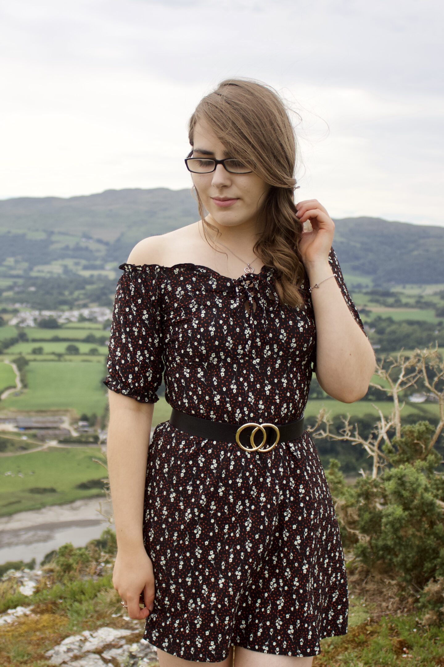 A photo of Elin wearing a black bardot floral mini dress, her hair is pulled over one shoulder and her left hand is raised, holding a strand away from her face
