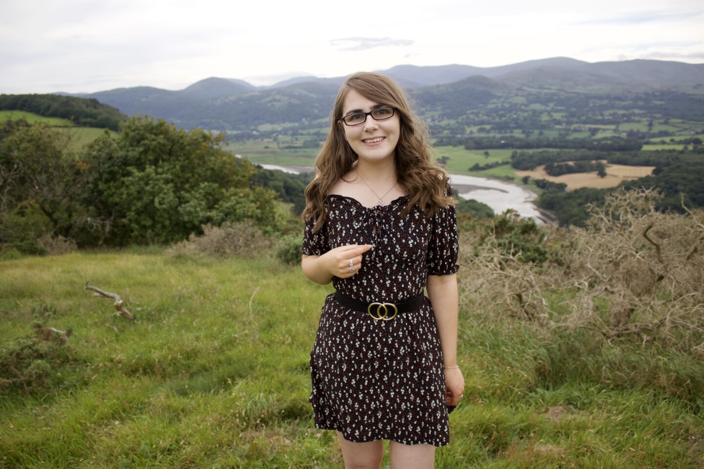A photo of Elin standing in front of a view of fields, a river and mountains, she wears a black floral mini dress and she's smiling at the camera