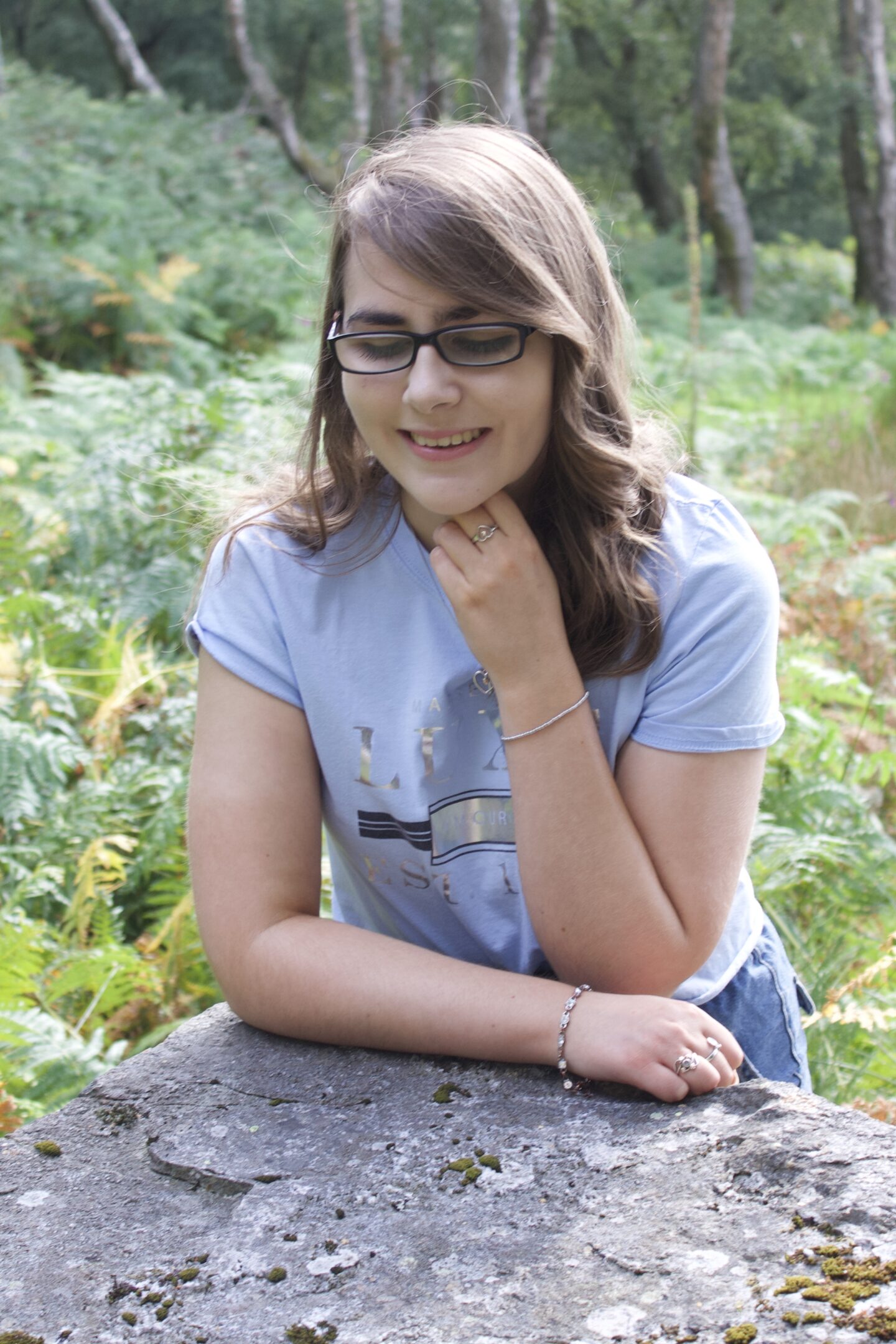 A photo of Elin leaning forward on a big rock, her right arm rests on the rock whilst her left is raised to underneath her chin. She's looking down and smiling