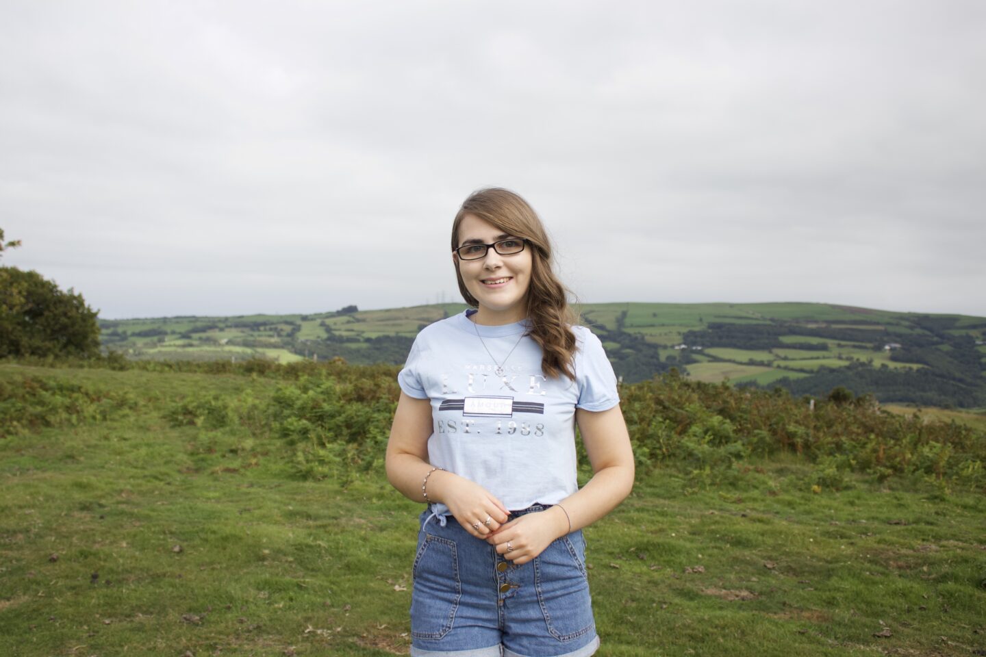 A photo of Elin standing in front of a view of fields, she's wearing a cropped light blue t-shirt with high waisted denim shorts. Her brown hair is curled