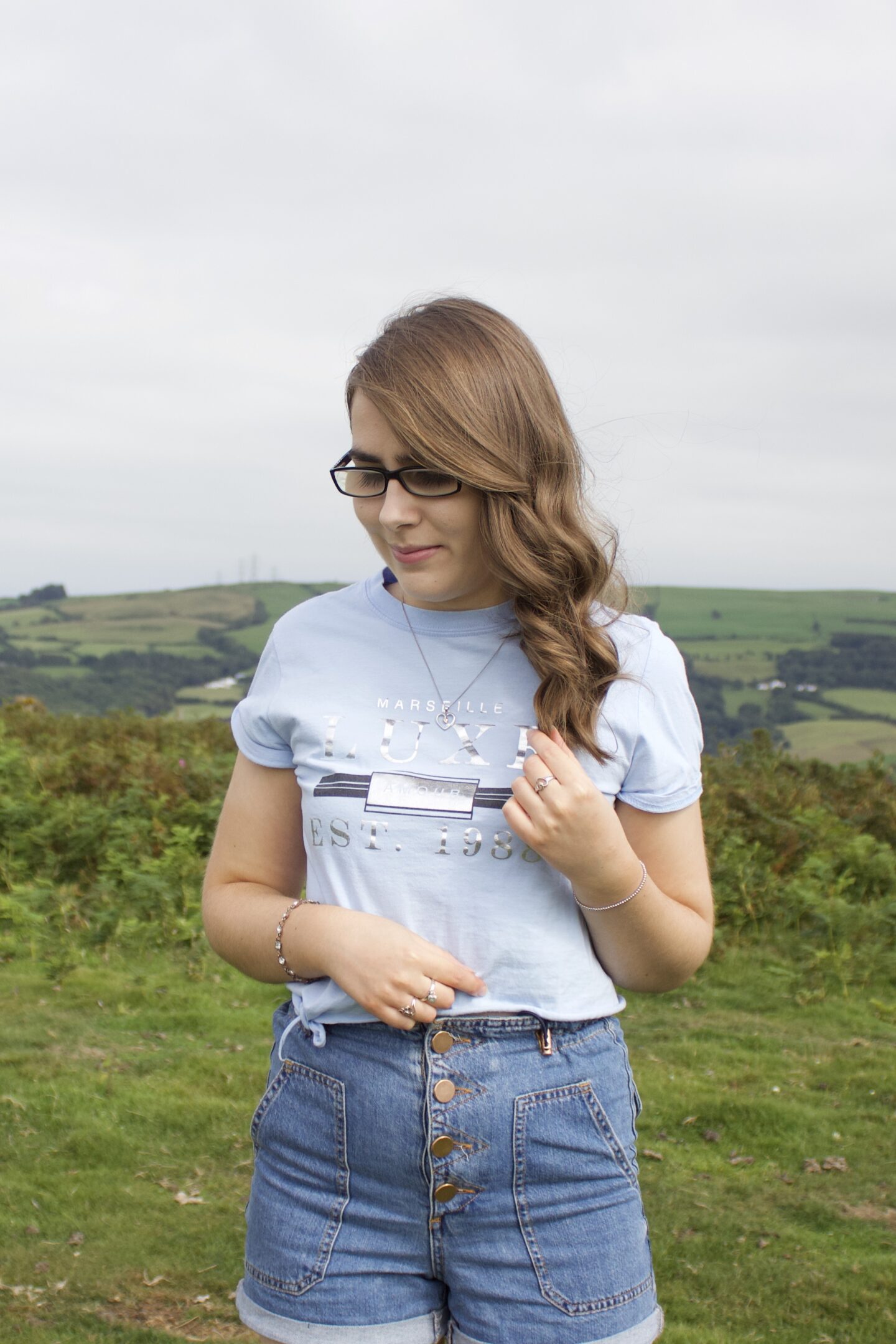 A photo of Elin standing in front of a view of fields, she's looking down and her left hand is raised, holding a strand of her curly hair