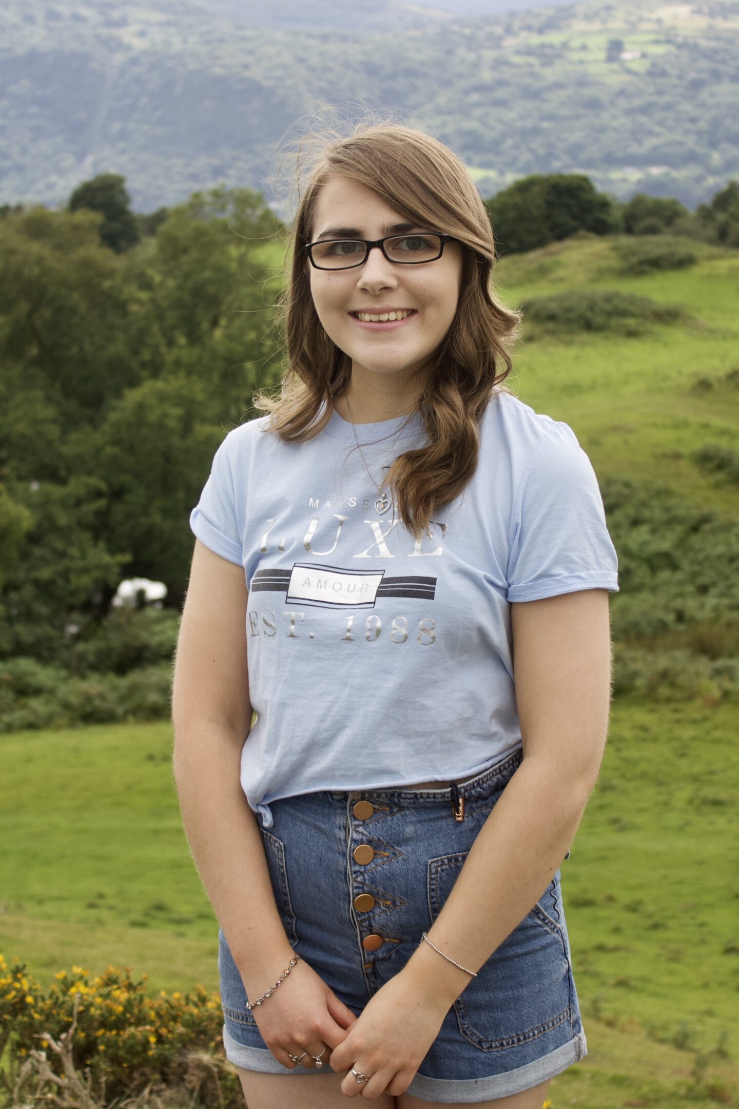 A photo of Elin standing in front of a view of mountains, she wears a light blue cropped t-shirt with high waisted denim shorts
