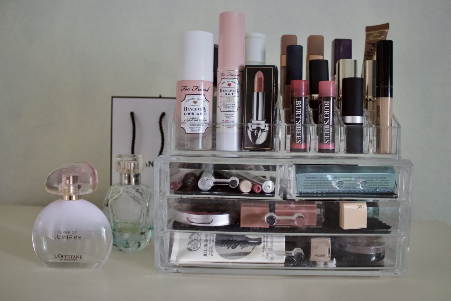 A clear makeup organiser which has a few drawers and a lipstick stand on top. There are a couple of perfumes to the left of the organiser, one is by L'occitane and it is in a clear and white bottle.