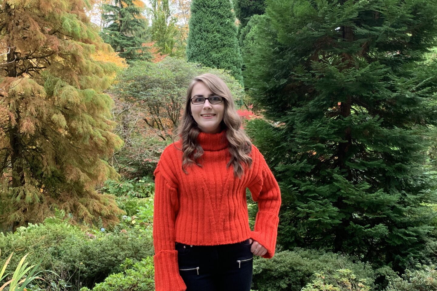 Elin standing in front of light orange coloured plants. She wears a red/orange knitted jumper with black skinny jeans. She's smiling at the camera and her left thumb is hooked inside her jeans pocket