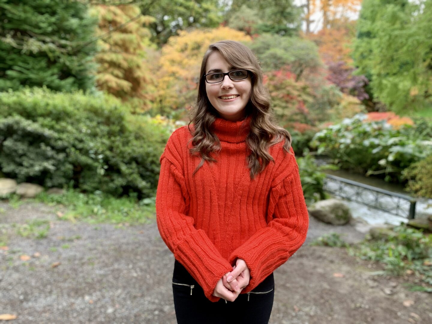 Elin standing in a wooded area, the background is blurred. She wears a red/orange knitted jumper and she's smiling at the camera. Her brown hair is curled.