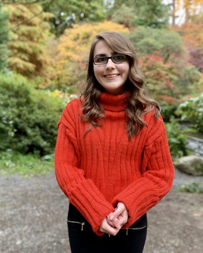 Elin standing in a wooded area, the background is blurred. She wears a red/orange knitted jumper and she's smiling at the camera. Her brown hair is curled.