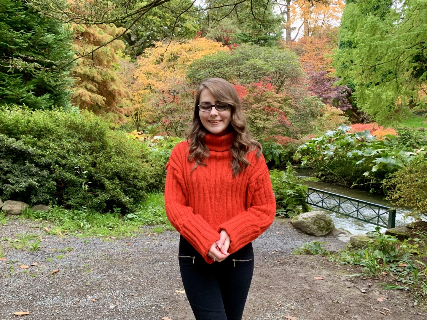 Elin standing in a wooded area with a river to her left, she's smiling and looking down