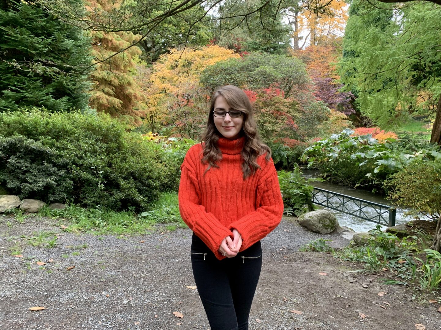 Elin wearing a red/orange cable knitted jumper which has a thick roll neck style, it's paired with black skinny jeans. She's looking down and there are yellow leafed trees to be seen behind her.