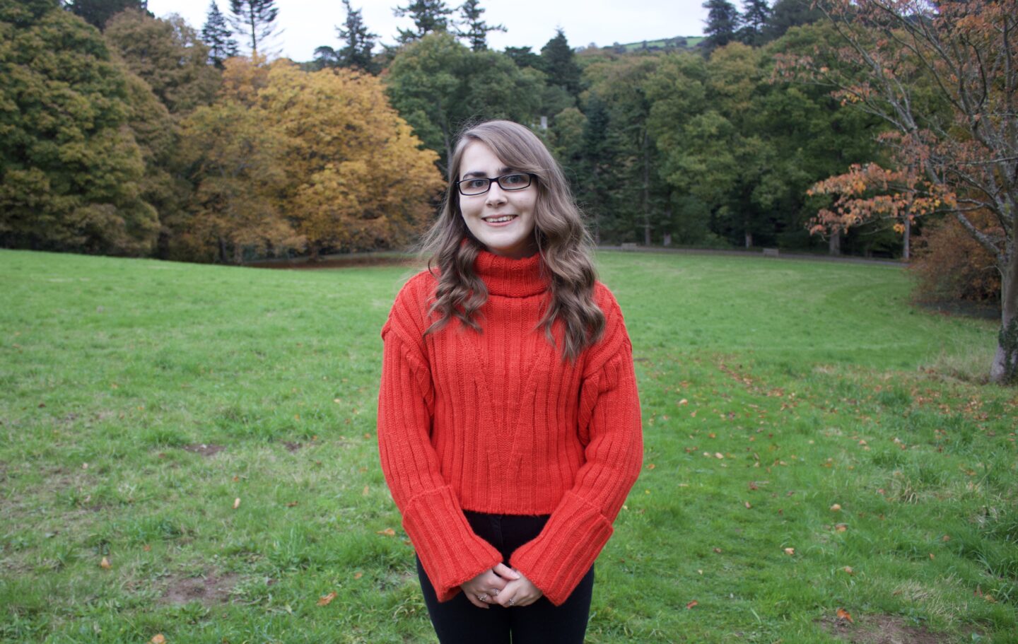 Elin standing in a field which is framed by pretty autumn coloured trees. She wears a red/orange knitted jumper with black jeans and she's smiling at the camera