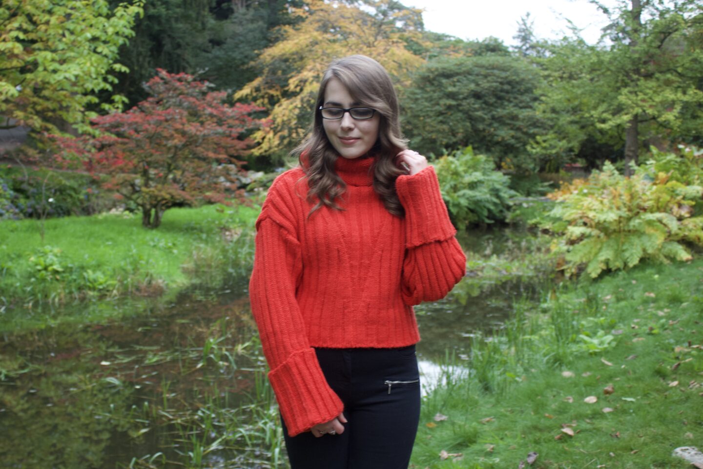 Elin standing in front of a small lake which has light yellow, orange and red coloured trees and plants behind it. Her left hand is holding a strand of her curly hair and she's looking down.