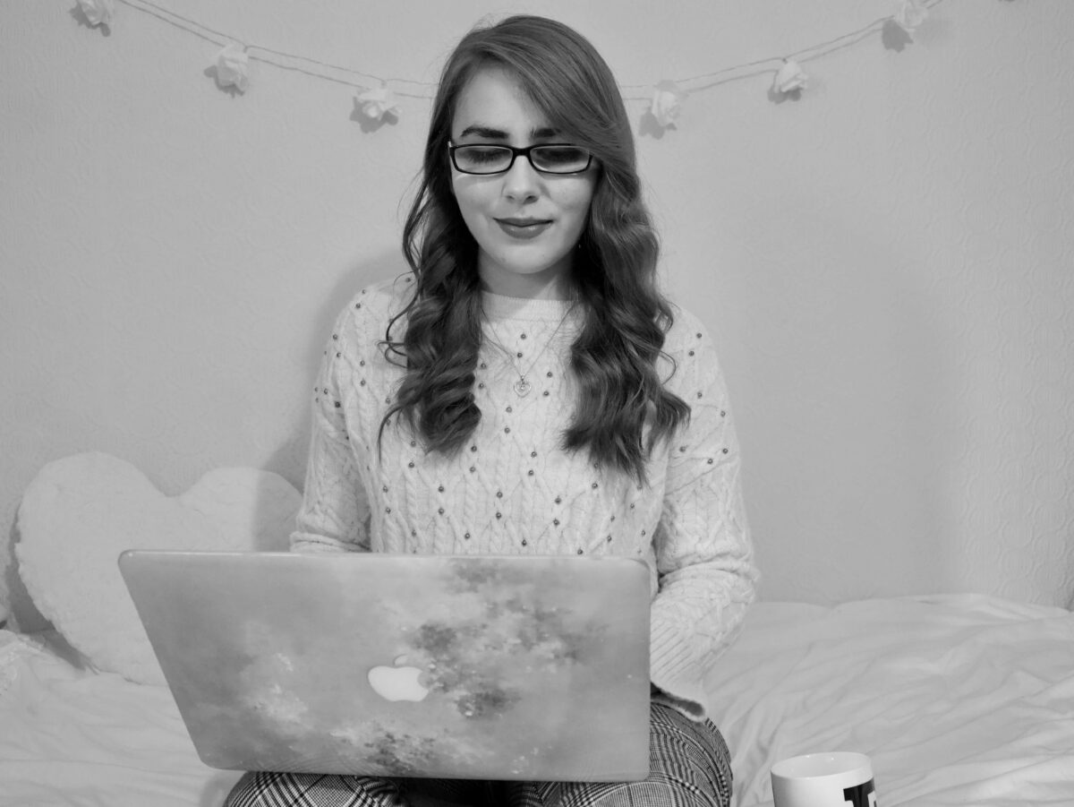 A black and white photo of Elin sitting on a bed with a laptop on her lap. She's looking at the laptop. There are floral shaped fairy lights on the wall behind her.