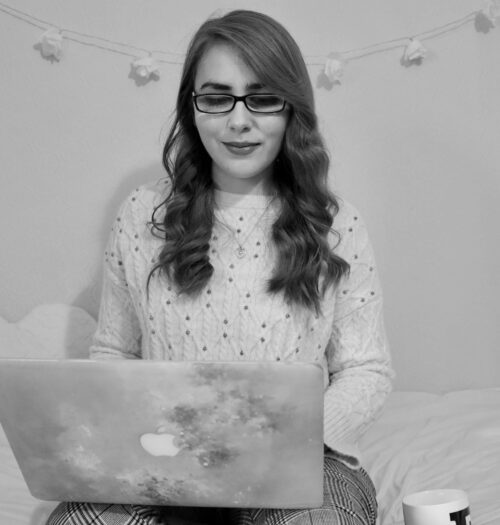 A black and white photo of Elin sitting on a bed with a laptop on her lap. She's looking at the laptop. There are floral shaped fairy lights on the wall behind her.
