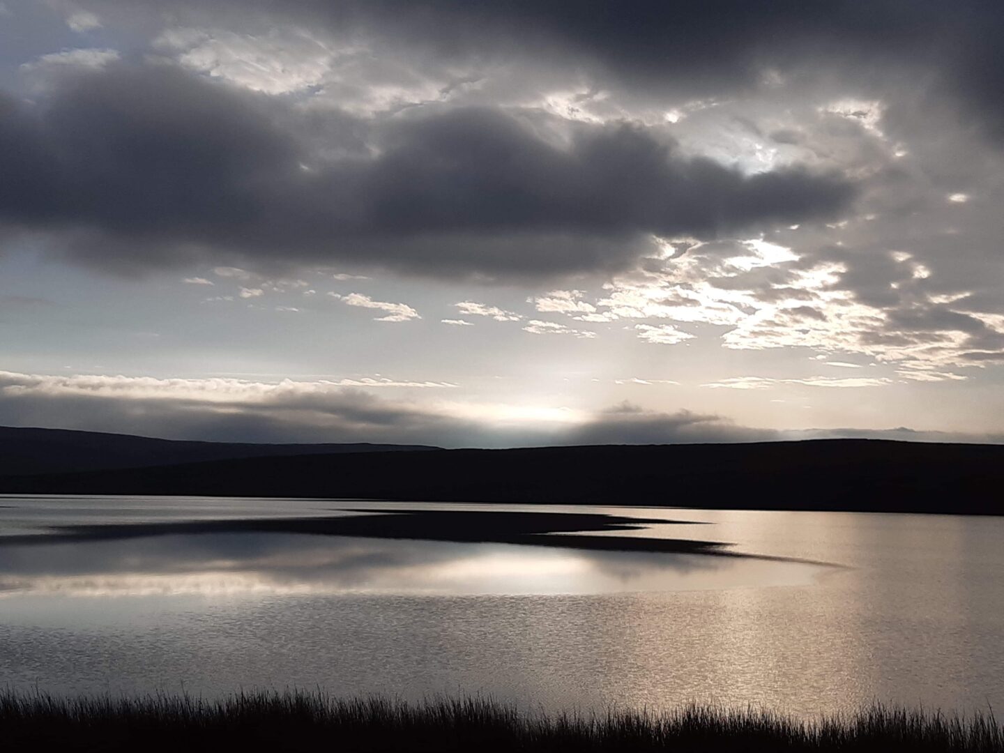 Grey clouds and the last of the day's sun reflecting on the calm water of a big lake which is surrounded by countryside