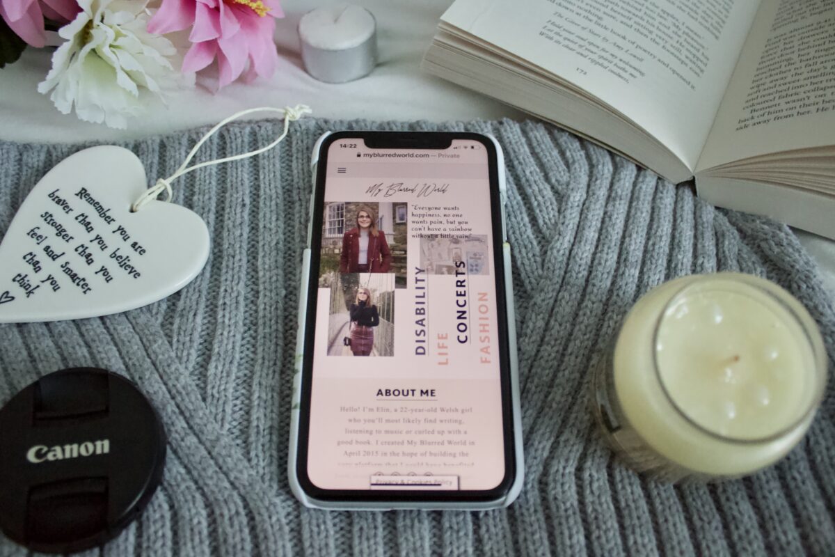 My Blurred World's homepage on an iPhone which is placed on a grey knitted scarf. The phone is surrounded by a white heart ornament, an open book and a candle