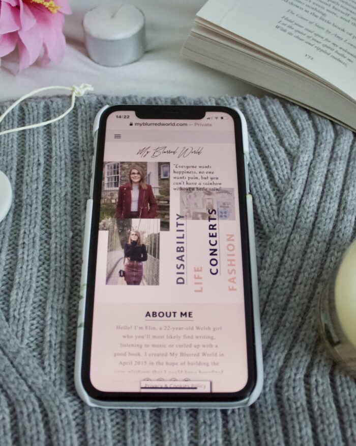 My Blurred World's homepage on an iPhone which is placed on a grey knitted scarf. The phone is surrounded by a white heart ornament, an open book and a candle