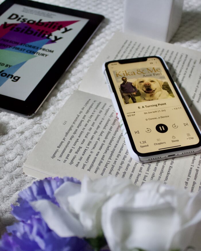 Fake blue and white flowers obscure the corner pages of an open book, an iPhone is placed on top of the book and the screen shows an audiobook version of Kika and Me by Dr Amit Patel
