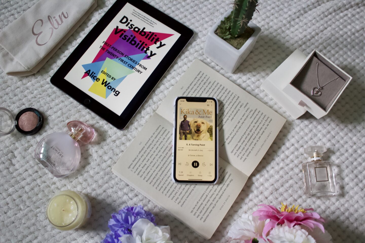 An audiobook version of Kika and Me by Dr Amit Patel is playing on an iPhone. The phone is placed on the pages of an open book. An iPad showing the front cover of Disability Visibility, edited by Alice Wong, and there are other items such as a candle and a couple of perfumes surrounding it