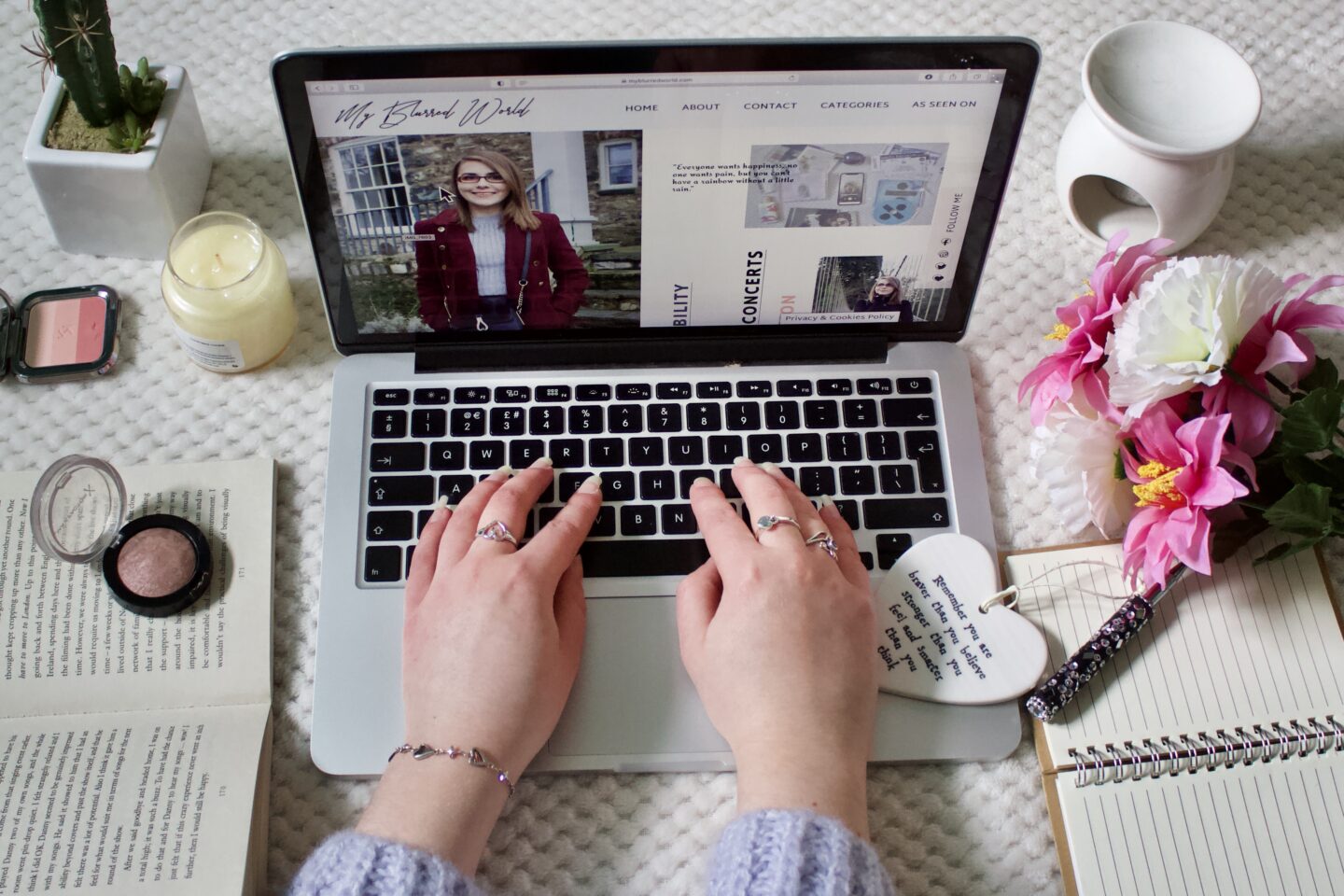 A laptop screen displaying My Blurred World’s homepage which shows a couple of photos of Elin. The laptop is place on a white textured blanket with various items arranged around it. To the left of the laptop, there is a small potted plant, an unlit candle and a couple of makeup items. To the right of the laptop, there is a white mug, a bouquet of pink artificial flowers, an open notebook with a pen on it and a heart-shaped ornament with text that is partially visible.
Elin’s hands are on the laptop keyboard with her lilac knitted jumper slightly visible. She wears three silver rings and a dainty bracelet with harps on it on her left wrist.