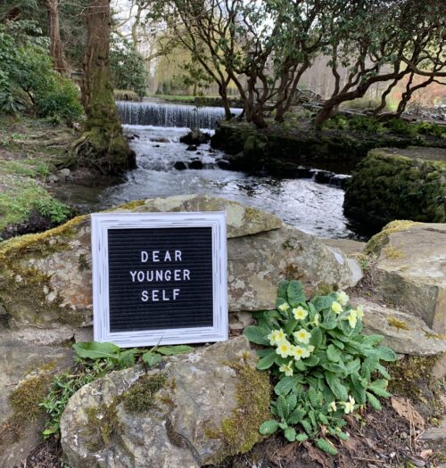 A black felt letter board with the words Dear Younger Self in white on it rests beside a primrose on some rocks in front of a river that is flanked by trees and plants