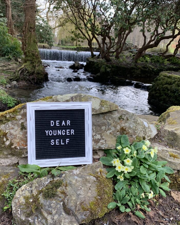 A black felt letter board with the words Dear Younger Self in white on it rests beside a primrose on some rocks in front of a river that is flanked by trees and plants