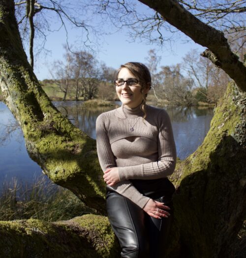 Elin sitting on a tree branch in front of a small lake which looks bright blue under the sun's glare. Elin and her surroundings are cast in shadows. She wears a beige high neck top which has a diamond shaped cutout, and black faux leather trousers. She's smiling and looking away from the camera to her right.