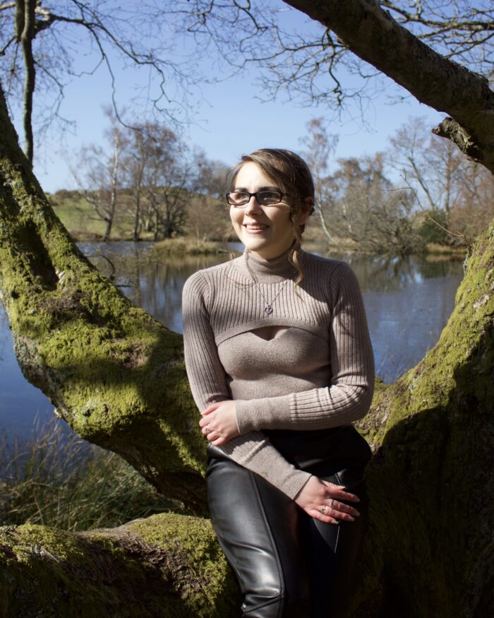 Elin sitting on a tree branch in front of a small lake which looks bright blue under the sun's glare. Elin and her surroundings are cast in shadows. She wears a beige high neck top which has a diamond shaped cutout, and black faux leather trousers. She's smiling and looking away from the camera to her right.