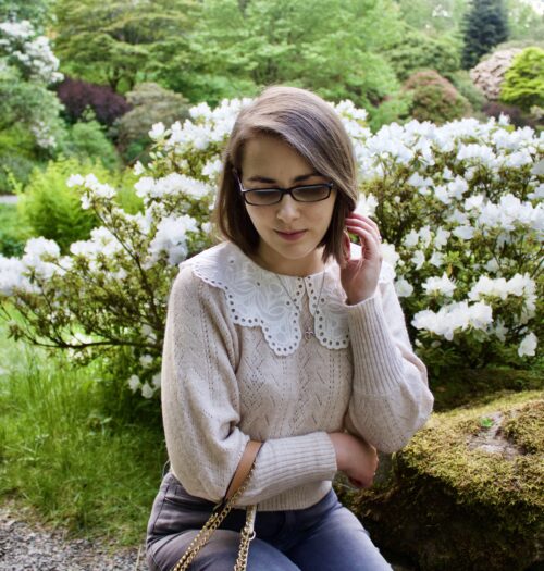 Elin crouching down in front of an array of white flowers. Her left hand is raised to push a strand of hair away from her face and she's looking down