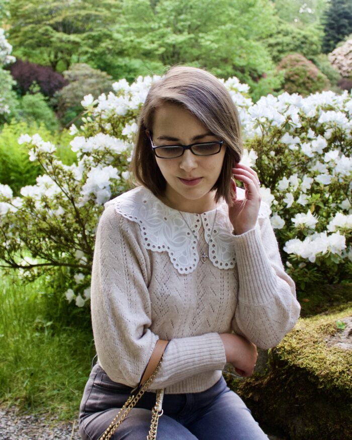 Elin crouching down in front of an array of white flowers. Her left hand is raised to push a strand of hair away from her face and she's looking down